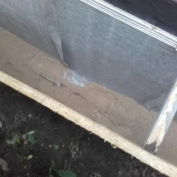 Wykop zabezpieczony folią, wypełniony piaskiem, widoczne drewniane szalunki i fragment buta pracownika.