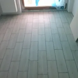 Świeżo ułożone, prostokątne, szare płytki podłogowe z plastikowymi krzyżykami dystansowymi w spoinach, widoczne przy oknie zabezpieczonym folią.