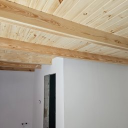KamAn - Drewniany sufit z belek w jasnym odcieniu, widoczny fragment ściany z otworem drzwiowym w surowym stanie. Ujęcie z dołu, podkreślające strukturę drewna.