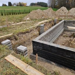 KamAn - Wykop pod fundamenty z częściowo wzniesionymi ścianami z bloczków betonowych, zabezpieczonymi czarną izolacją. W tle ziemia i zielone wzgórza.