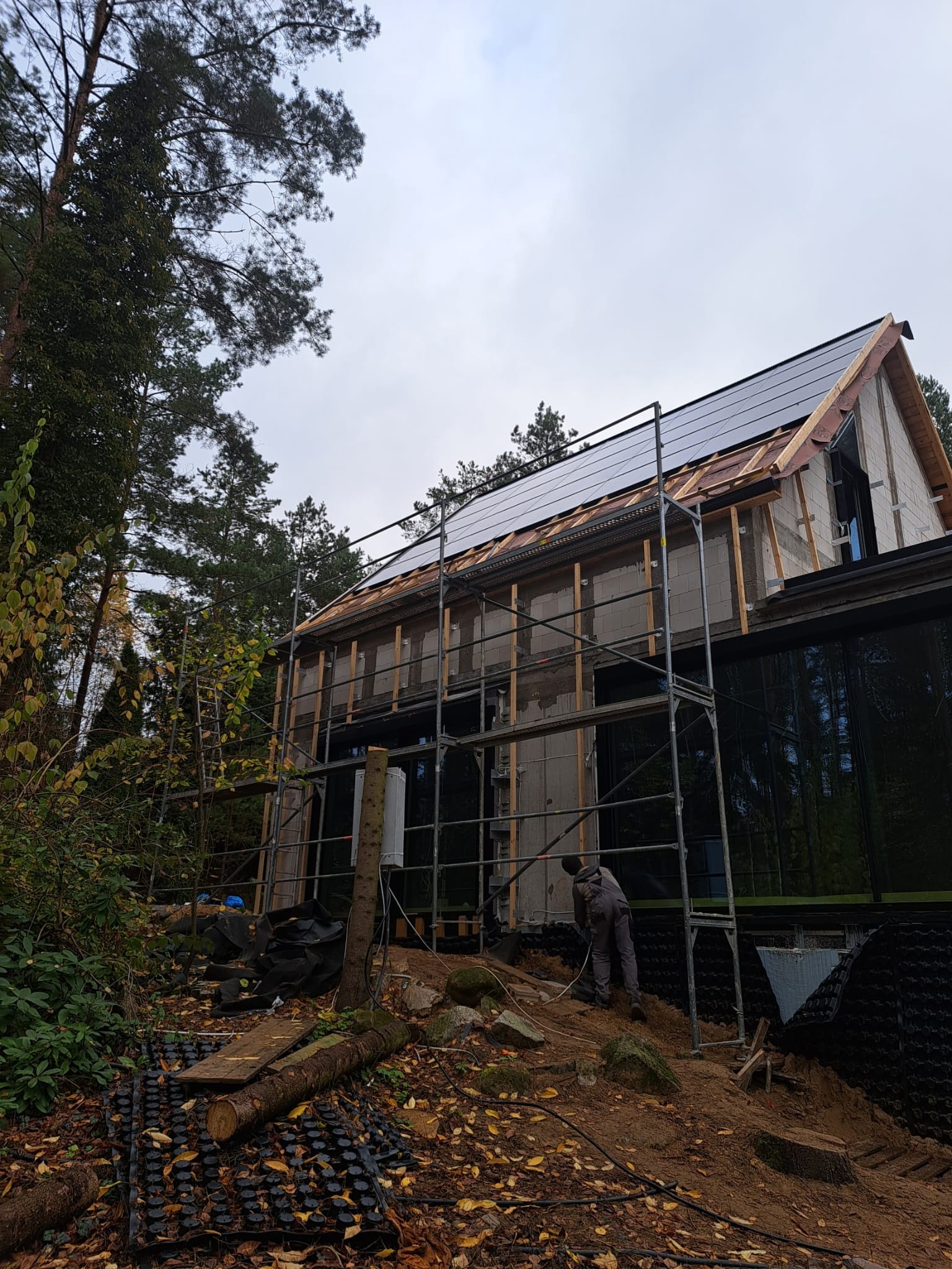 Budowa domu w lesie: pracownik na rusztowaniu montuje panele słoneczne na dachu. Widoczne bloczki konstrukcyjne, okna i elementy izolacji. Otoczenie leśne.