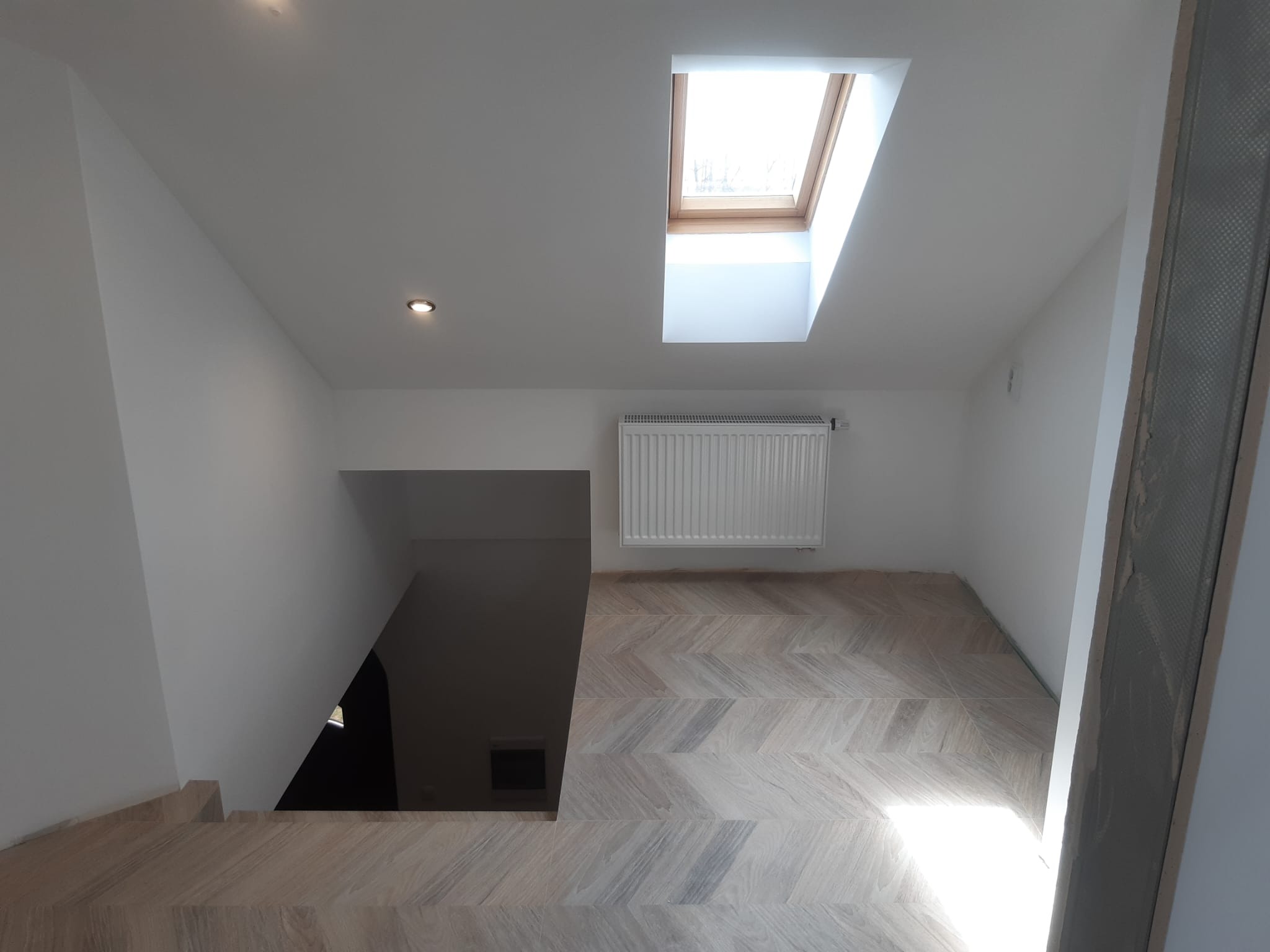 Jasne wnętrze z drewnianą podłogą ułożoną w jodełkę, schody w lewo, okno dachowe wpuszczające światło. Widok z góry na wykończone wnętrze.