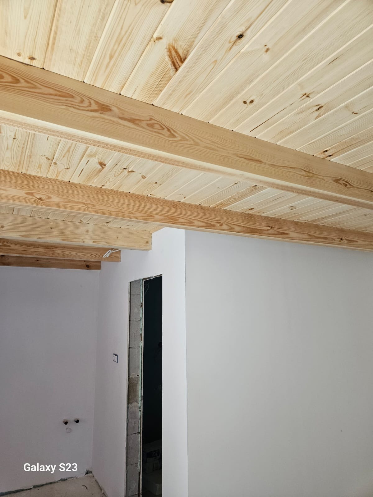 Drewniany sufit z belek w jasnym odcieniu, widoczny fragment ściany z otworem drzwiowym w surowym stanie. Ujęcie z dołu, podkreślające strukturę drewna.