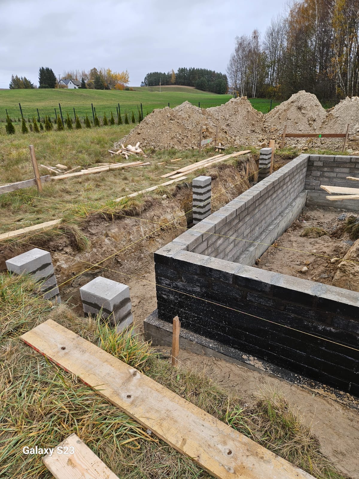 Wykop pod fundamenty z częściowo wzniesionymi ścianami z bloczków betonowych, zabezpieczonymi czarną izolacją. W tle ziemia i zielone wzgórza.