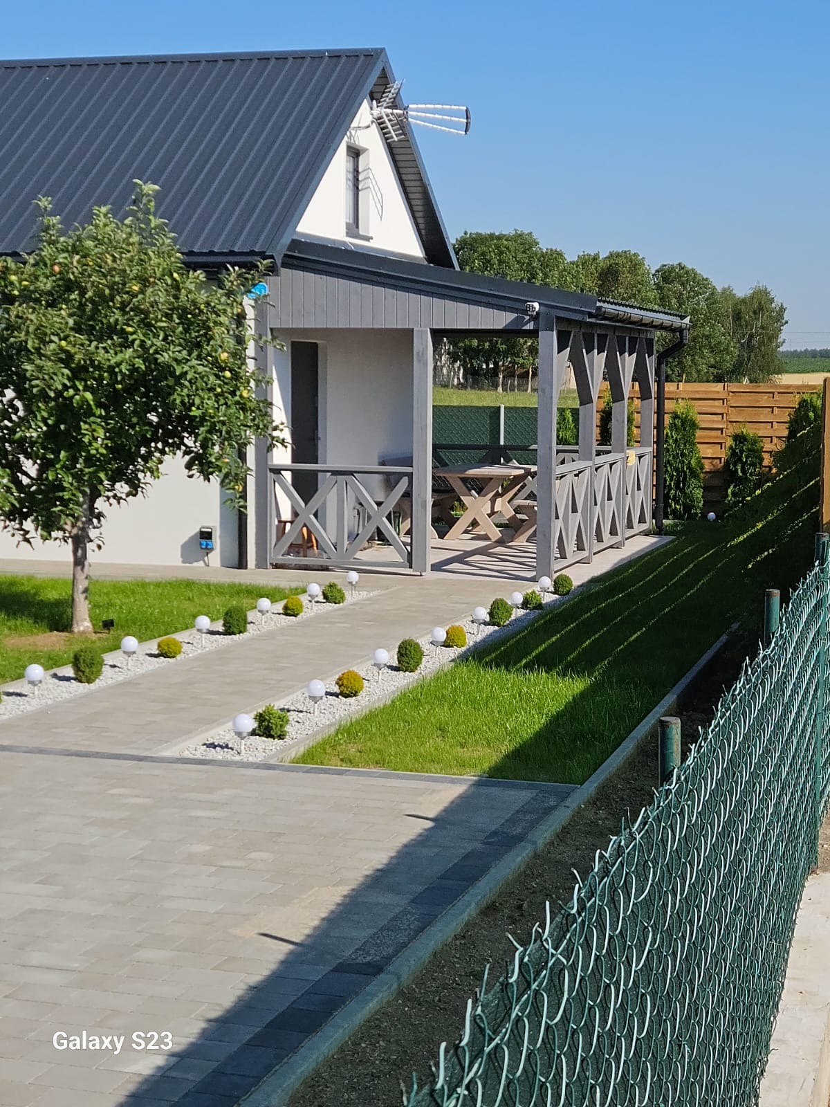 Nowoczesny dom z szarą altaną i zielonym ogrodem. Ścieżka z kostki prowadzi przez trawnik z kulistymi krzewami i lampami. W tle widoczne zielone ogrodzenie z siatki.