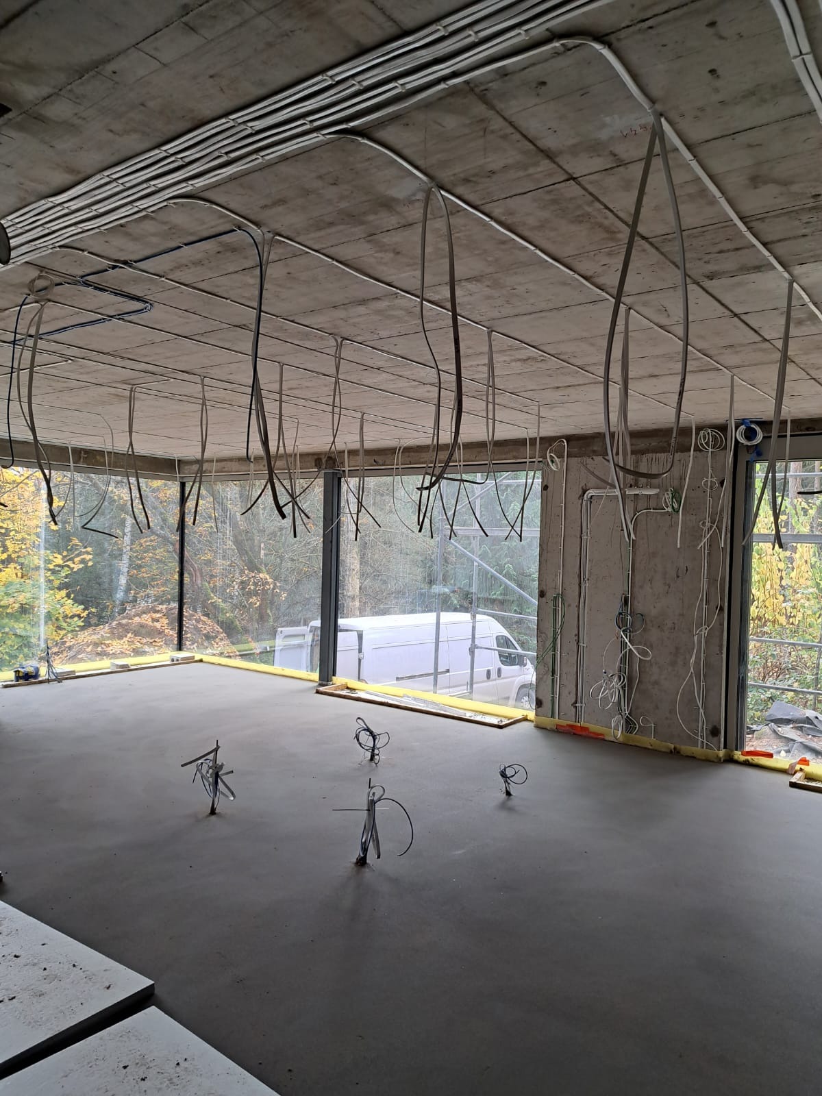 Surowy stan wnętrza z widoczną instalacją elektryczną na suficie i w ścianach, przygotowany do dalszych prac wykończeniowych. Duże okna z widokiem na zieleń.