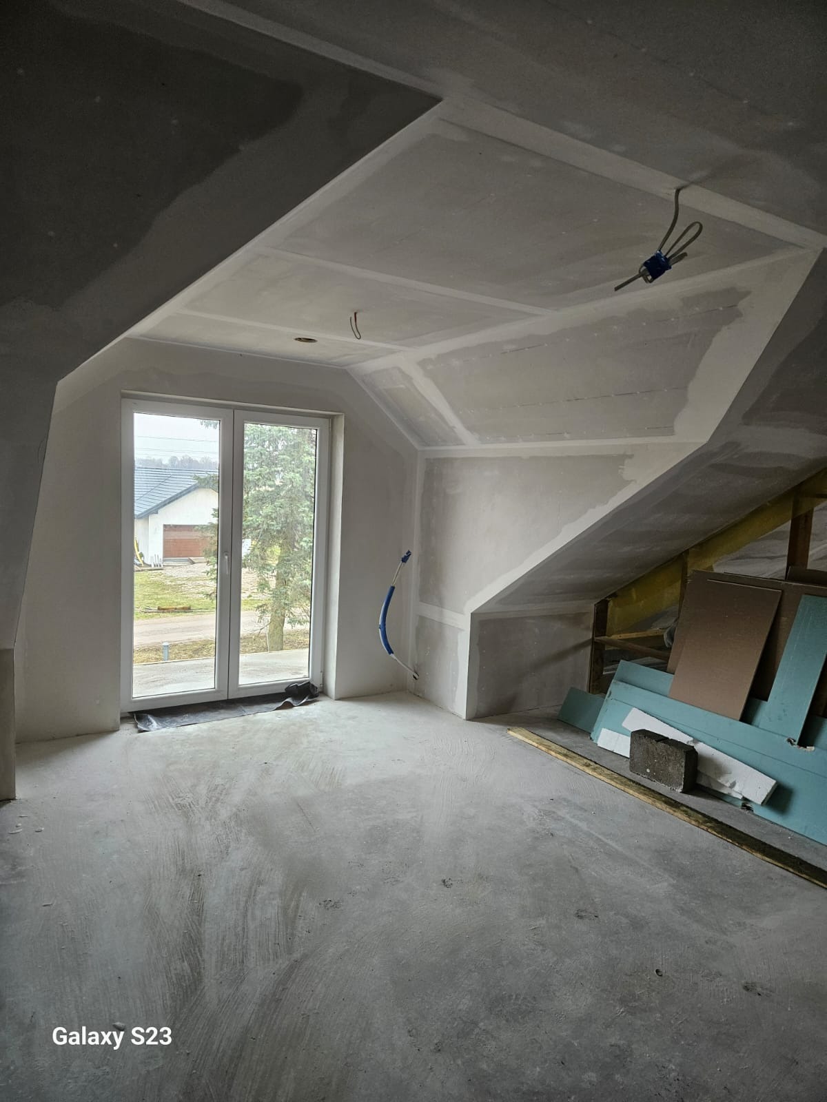 Poddasze w trakcie wykańczania: widoczne płyty gipsowo-kartonowe na skosach, surowa podłoga, okno balkonowe i materiały budowlane. W tle dom.