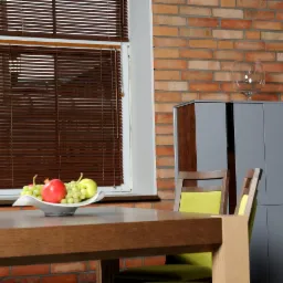 Brązowe poziome osłony okienne w pokoju z ceglaną ścianą, owocami na stole i nowoczesnymi szafkami w tle.