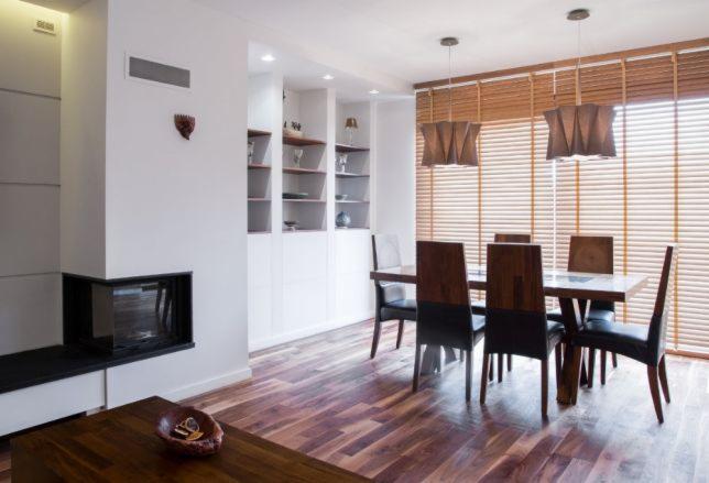 Jasne wnętrze jadalni z drewnianą podłogą, białymi ścianami, kominkiem, zabudowaną szafą i oknami przesłoniętymi poziomymi osłonami okiennymi w odcieniach brązu, z wiszącymi lampami o geometrycznym...