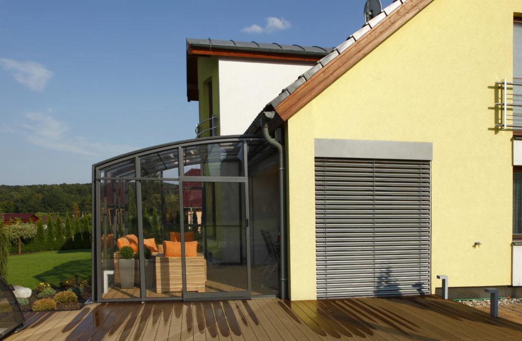 Żółty dom z przeszkloną werandą i szarymi zewnętrznymi osłonami okiennymi, widoczne odbicie na mokrym drewnianym tarasie.
