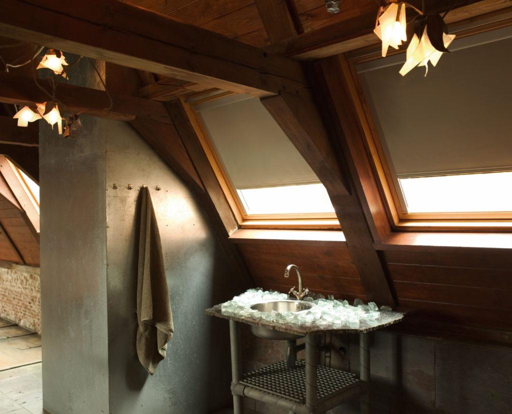 Poddasze z drewnianą konstrukcją, okna dachowe z zasłoniętymi roletami, umywalka z blatem z potłuczonego szkła i wiszący ręcznik.