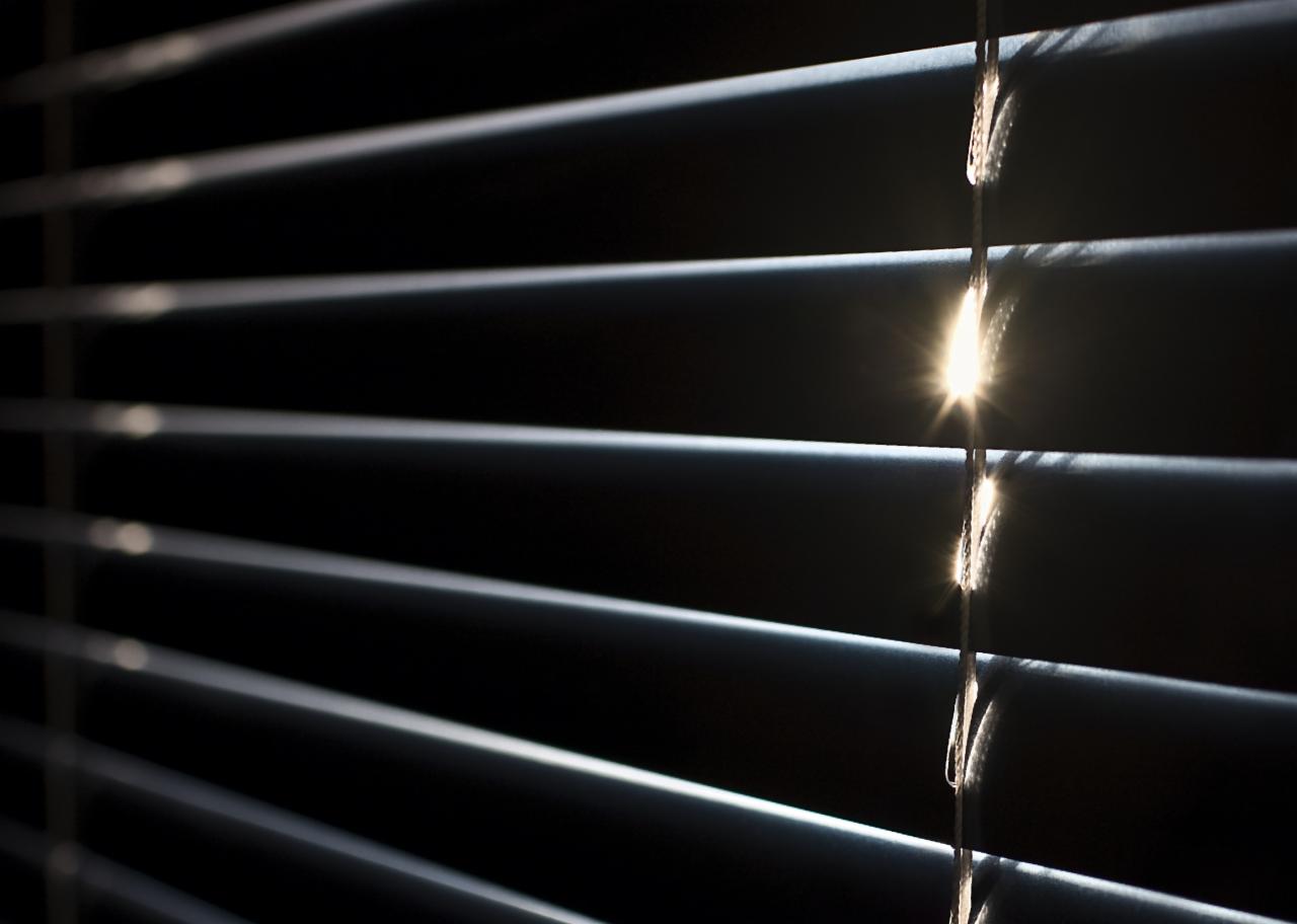 Ciemne poziome listwy z delikatnym prześwitem słońca, tworzące kontrastowe światło na sznurku sterującym.