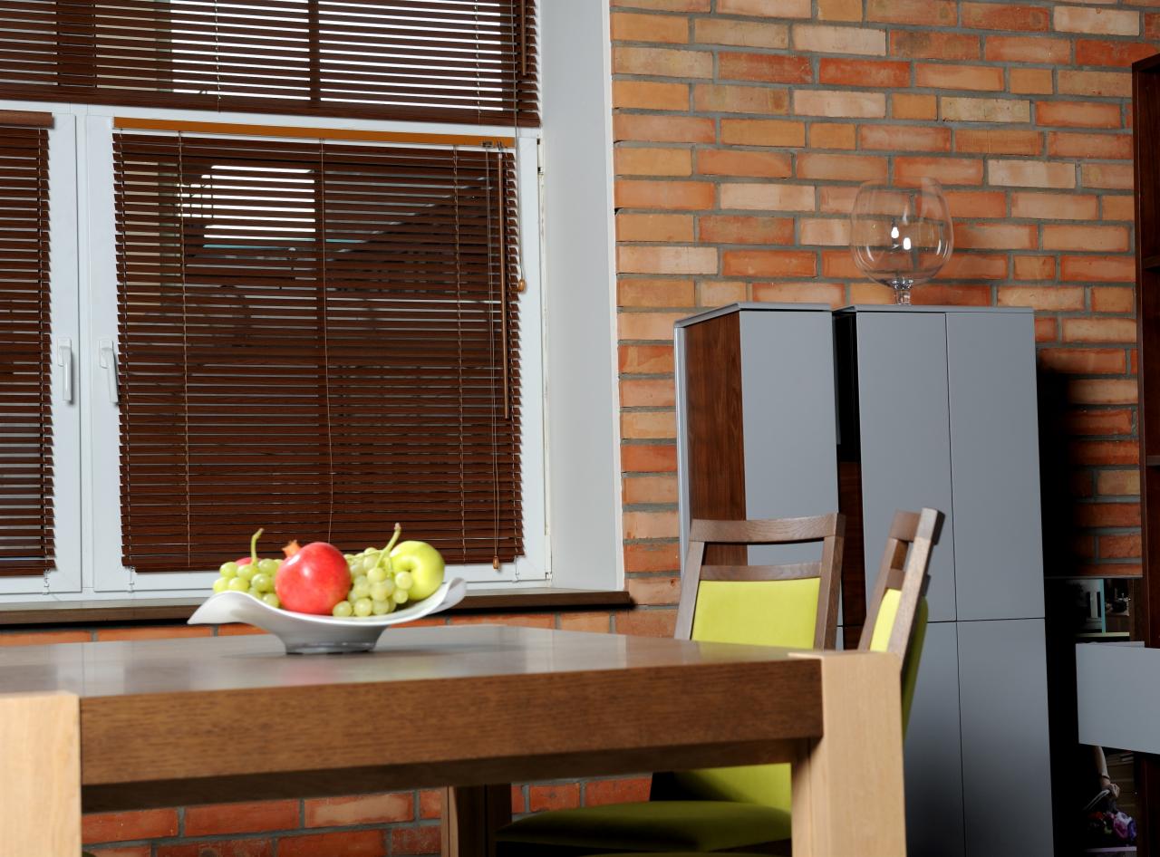 Brązowe poziome osłony okienne w pokoju z ceglaną ścianą, owocami na stole i nowoczesnymi szafkami w tle.