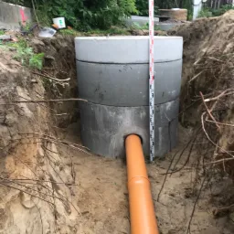 Betonowy zbiornik z podłączoną pomarańczową rurą kanalizacyjną, umieszczony w wykopie ziemnym. W tle widoczny niwelator laserowy i roślinność.