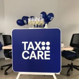 Stół z logo Tax Care, szampanem i balonami w biurze, krzesła biurowe po bokach, roślina w tle po lewej.