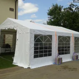 Biały namiot plenerowy z oknami łukowymi, zabezpieczony obciążnikami z cegieł na niebieskich podkładkach, ustawiony na kostce brukowej przed budynkiem z numerem 17.