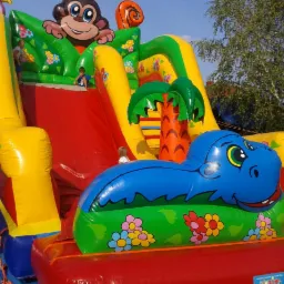 Kolorowy dmuchany zamek ze zjeżdżalnią, motyw małpy na szczycie, z boku niebieski hipopotam, dzieci na konstrukcji.