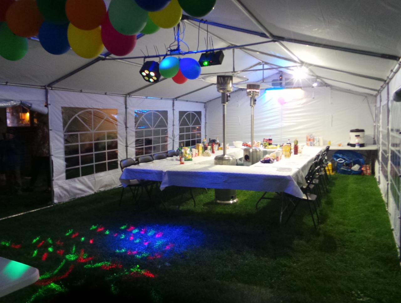 Wnętrze białego namiotu imprezowego z oświetleniem dyskotekowym na trawie, stołem nakrytym białym obrusem, krzesłami, dekoracjami i balonami pod sufitem.