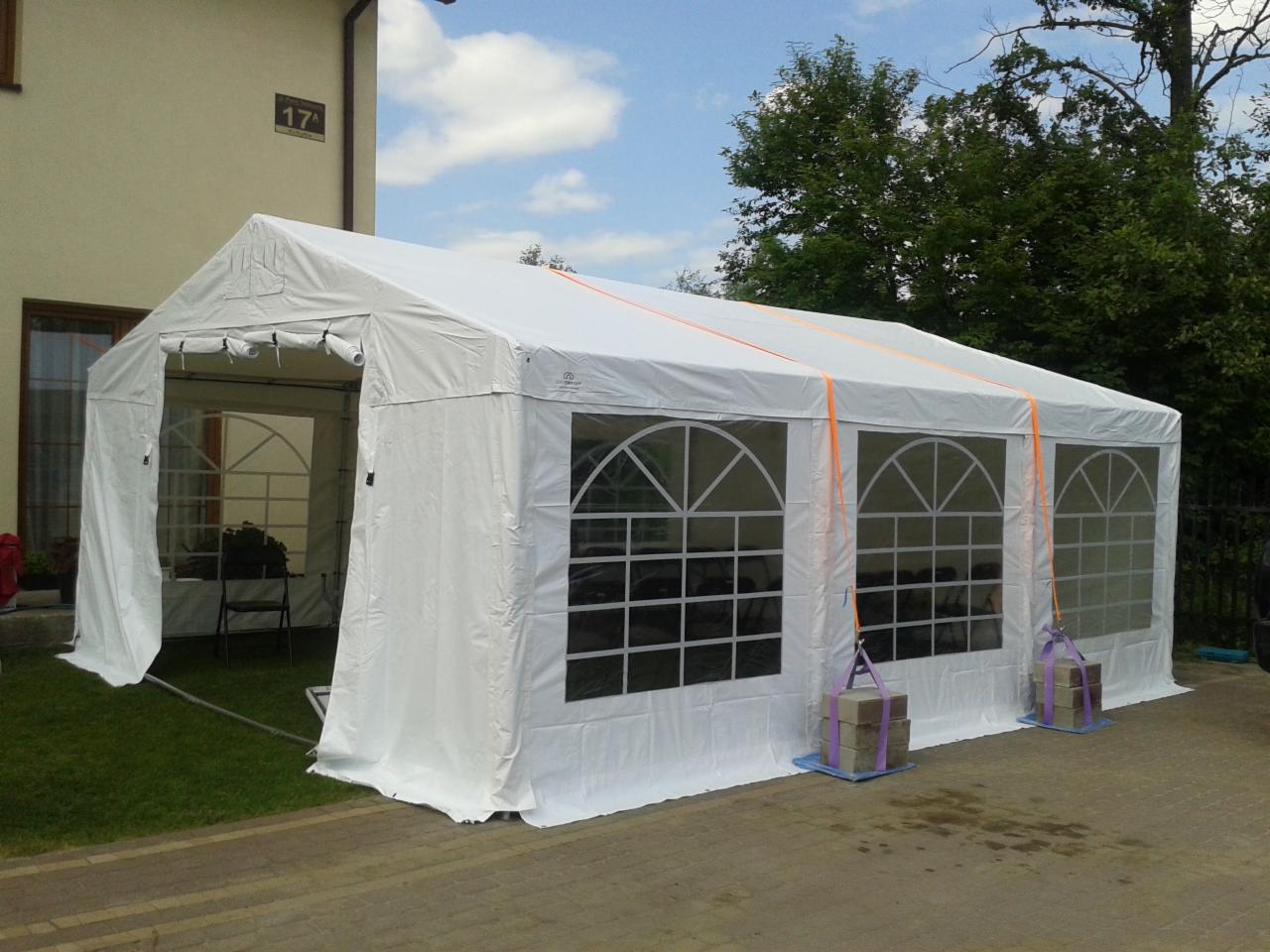 Biały namiot plenerowy z oknami łukowymi, zabezpieczony obciążnikami z cegieł na niebieskich podkładkach, ustawiony na kostce brukowej przed budynkiem z numerem 17.