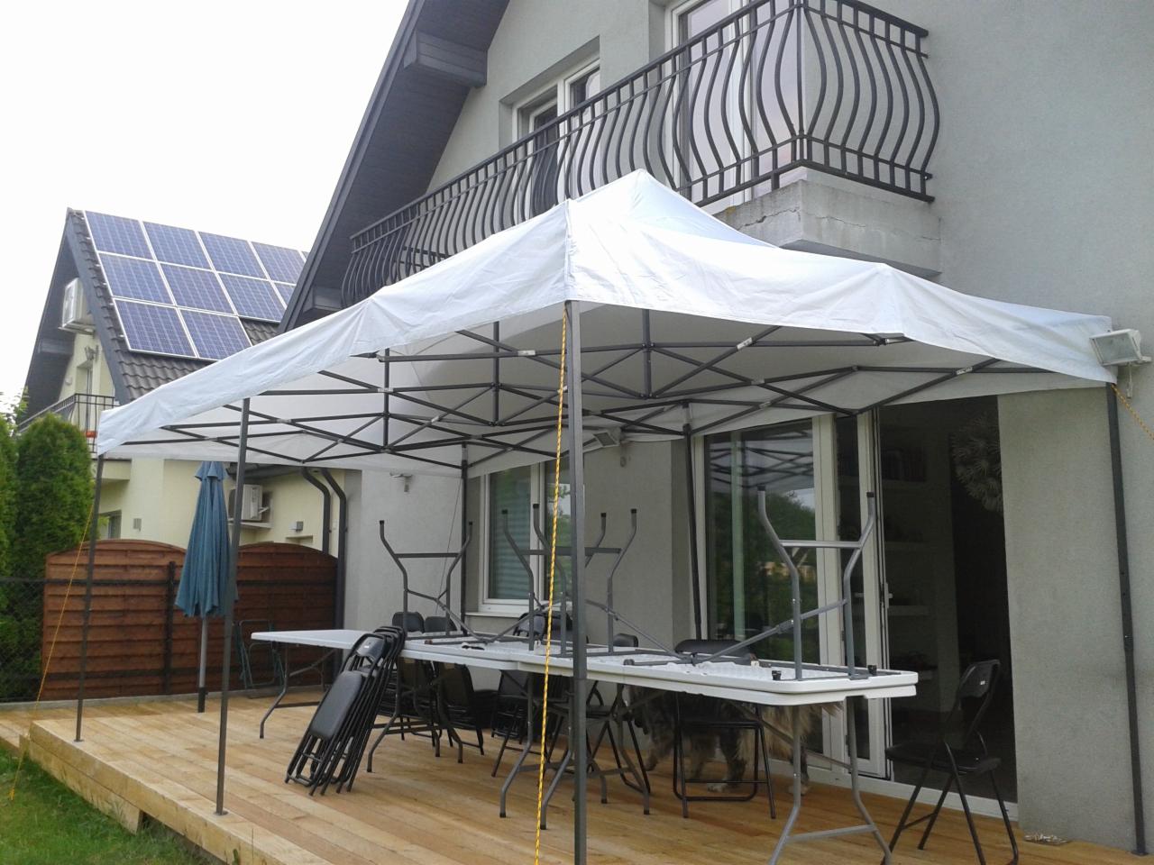 Rozłożony biały namiot plenerowy na drewnianym tarasie, z przygotowanymi stołami i krzesłami, widok na dom z balkonem i panele słoneczne na dachu sąsiedniego budynku.