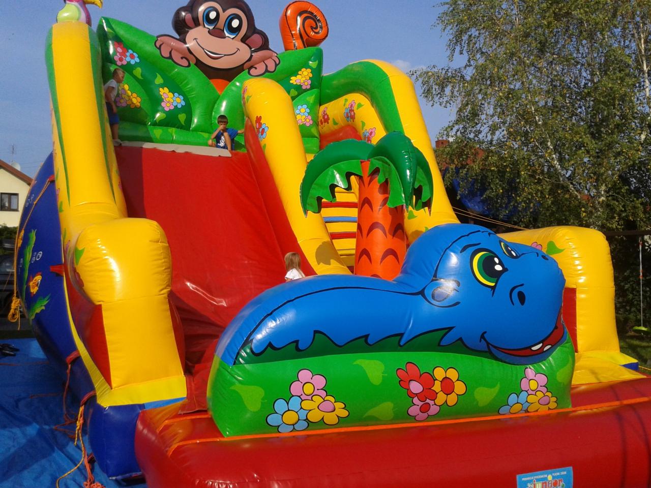 Kolorowy dmuchany zamek ze zjeżdżalnią, motyw małpy na szczycie, z boku niebieski hipopotam, dzieci na konstrukcji.
