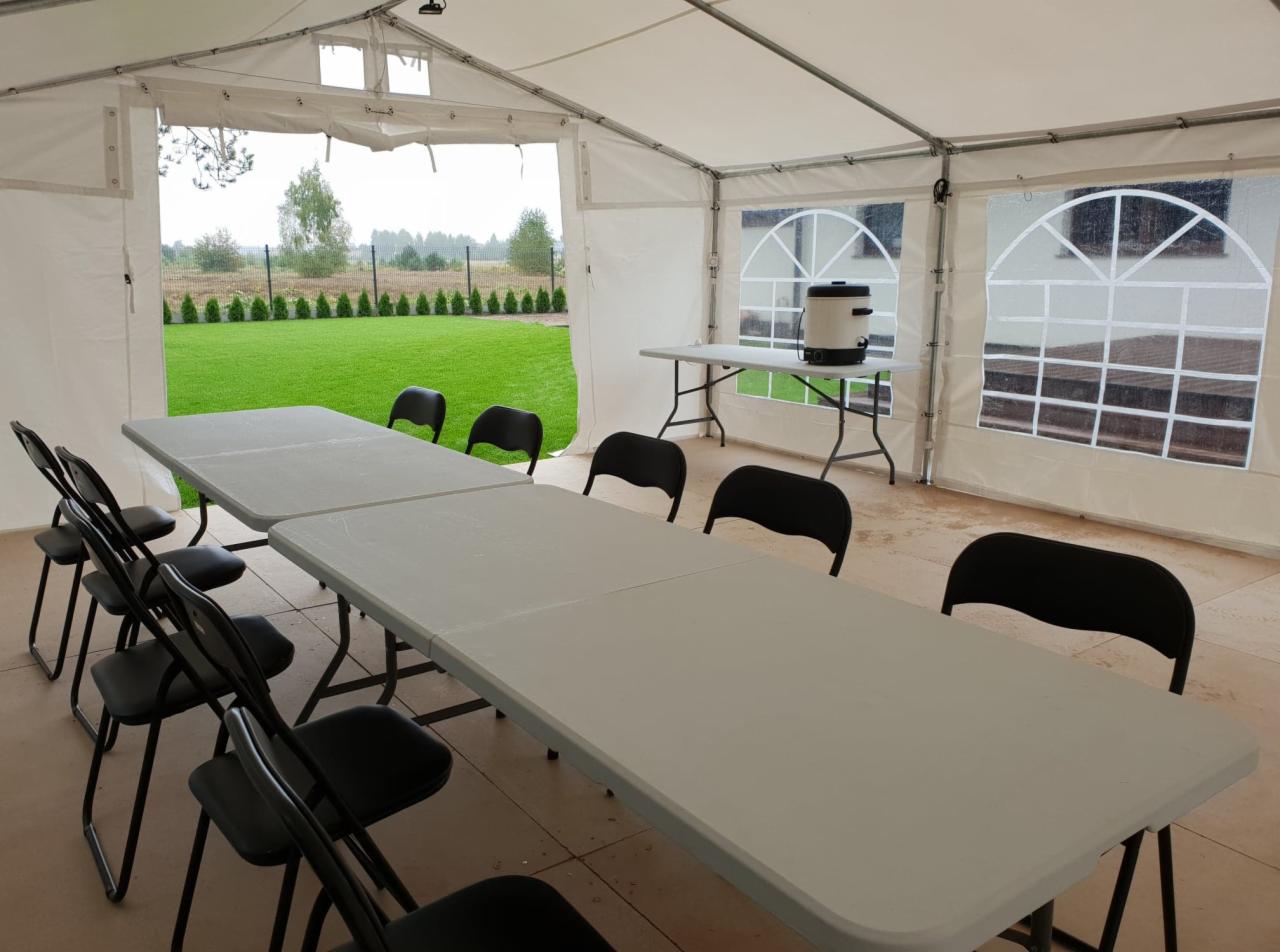 Wnętrze białego namiotu imprezowego z rozstawionymi stołami i krzesłami, widok na zewnątrz na trawnik i drzewa przez otwarte wejście i okna.