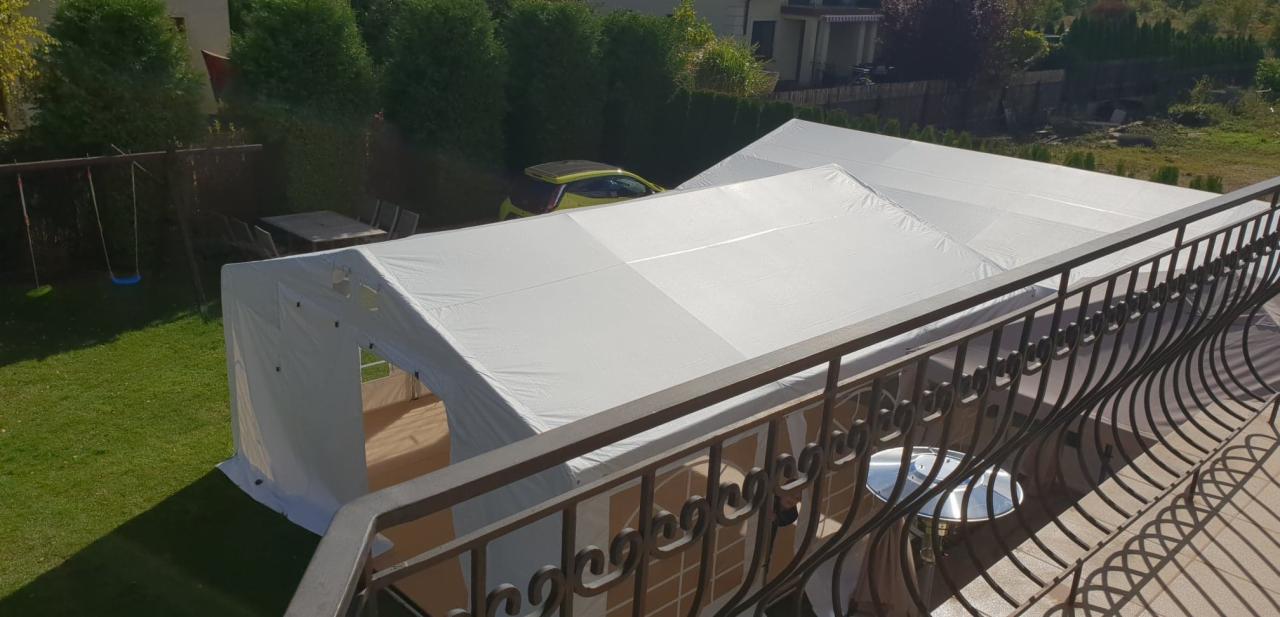 Dwa białe namioty plenerowe rozstawione na trawniku, widok z balkonu z ozdobną balustradą, w tle zieleń i żółty samochód.