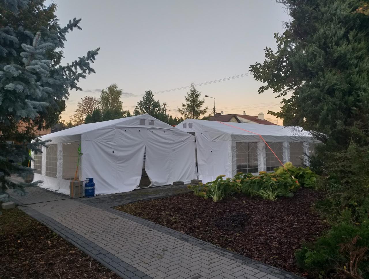 Dwa białe namioty połączone ze sobą, ustawione na zewnątrz na trawniku, widoczne drzewa i krzewy, niebieska butla z gazem przy jednym z namiotów.