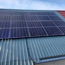 Panele fotowoltaiczne zamontowane na dachu z blachy trapezowej, widoczny kabel biegnący po dachu oraz fragment dachu z czerwonej dachówki w tle.