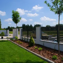 Nowoczesne ogrodzenie panelowe z poziomymi przęsłami w kolorze grafitowym, osadzone na białych słupkach, oddzielające trawnik od rabaty z korą i młodymi roślinami. W tle widoczny czerwony samochód...