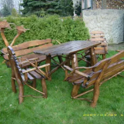 Rustykalny komplet mebli ogrodowych z litego drewna, składający się z prostokątnego stołu i kilku krzeseł/ławek, ustawiony na trawniku przed żywopłotem i betonowym murem.