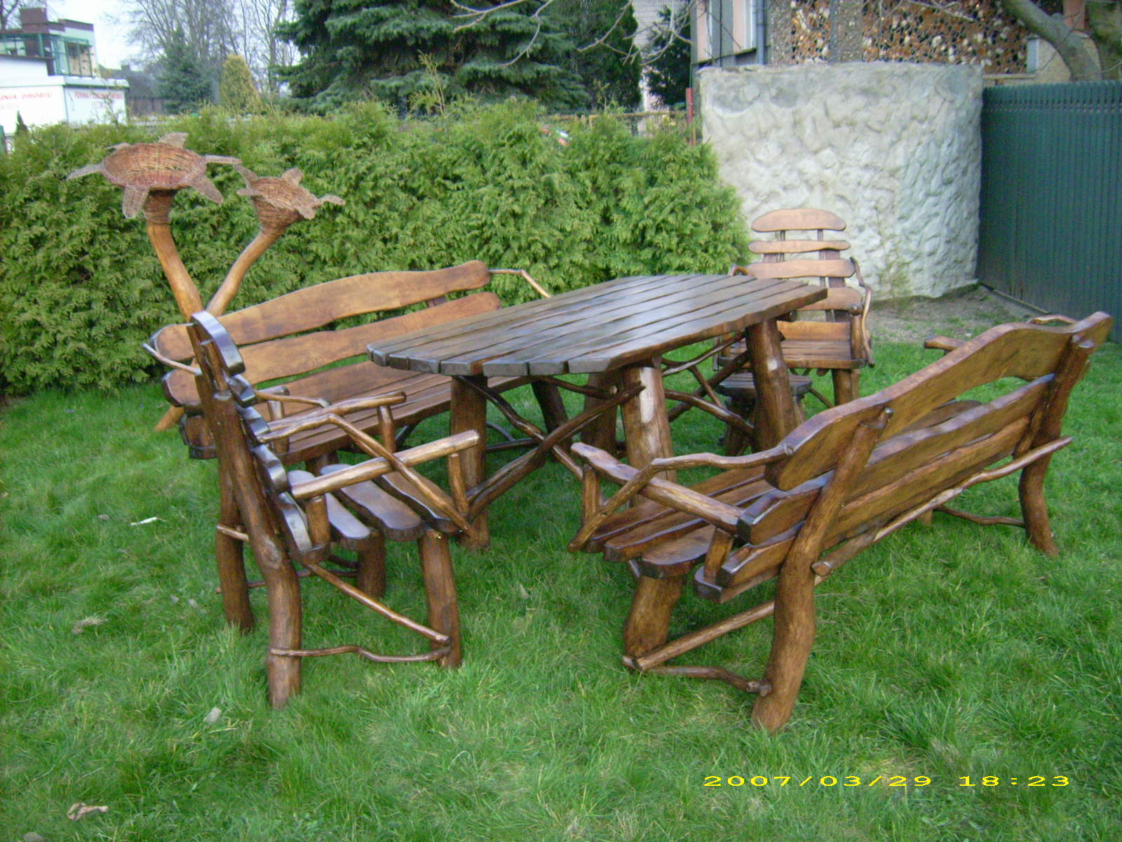 Rustykalny komplet mebli ogrodowych z litego drewna, składający się z prostokątnego stołu i kilku krzeseł/ławek, ustawiony na trawniku przed żywopłotem i betonowym murem.