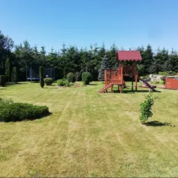 Rozległy trawnik z widocznymi śladami koszenia, otoczony różnorodną roślinnością: drzewami iglastymi, krzewami ozdobnymi o regularnych kształtach oraz trampoliną i drewnianym placem zabaw.