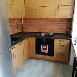 Jasna kuchnia z drewnianymi frontami, czarnym blatem i ceglaną ścianą nad blatem. Widoczna płyta grzewcza i piekarnik w zabudowie.