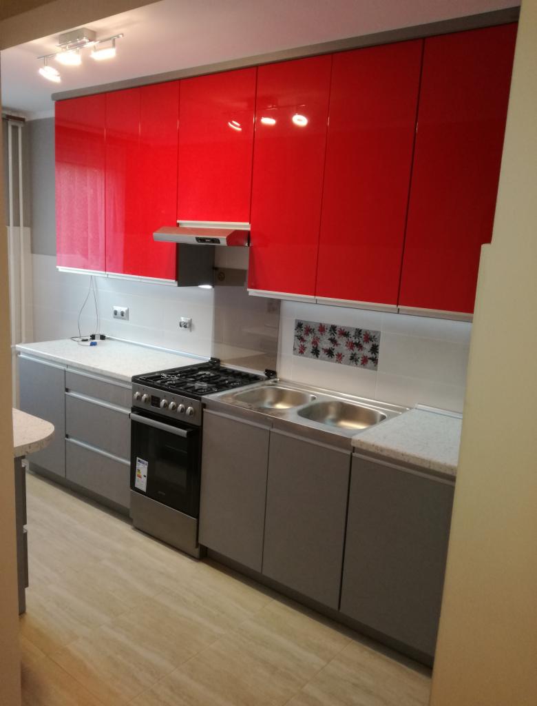 Nowoczesna kuchnia z czerwonymi szafkami górnymi na wysoki połysk i szarymi szafkami dolnymi, dwukomorowy zlewozmywak ze stali nierdzewnej, płyta gazowa i piekarnik, jasna podłoga imitująca drewno.
