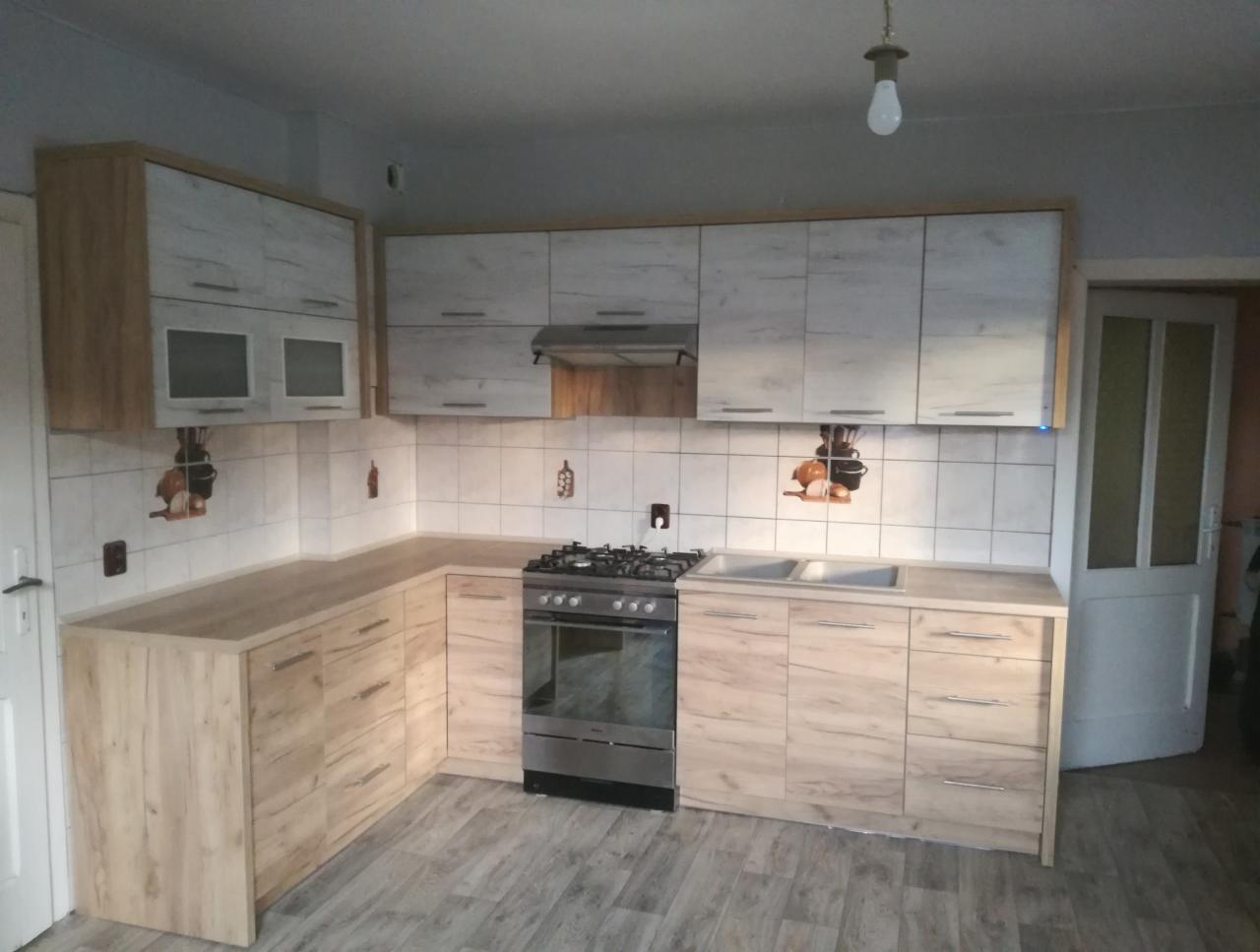 Jasna kuchnia w kształcie litery L z drewnopodobnymi frontami, białymi kafelkami na ścianie i kuchenką gazową ze stali nierdzewnej.