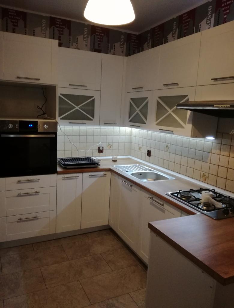 Biała zabudowa kuchenna w kształcie litery L z przeszklonymi szafkami górnymi z motywem krzyża, oświetleniem podszafkowym i drewnianym blatem, widok na płytę gazową i zlewozmywak.