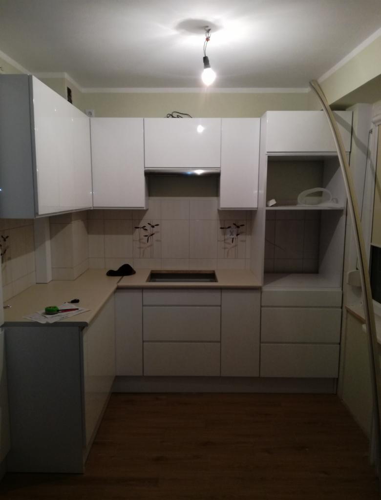 Jasna kuchnia z białymi frontami szafek, widoczne akcesoria na blacie i płytki z delikatnym wzorem na ścianie.