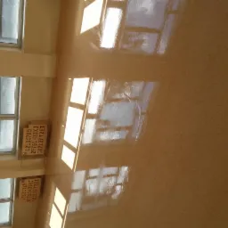 Błyszcząca, świeżo umyta podłoga w jasnym pomieszczeniu z oknami, na której widoczne są refleksy światła słonecznego i niewyraźne ślady po czyszczeniu. W rogu pomieszczenia widoczne kartony...