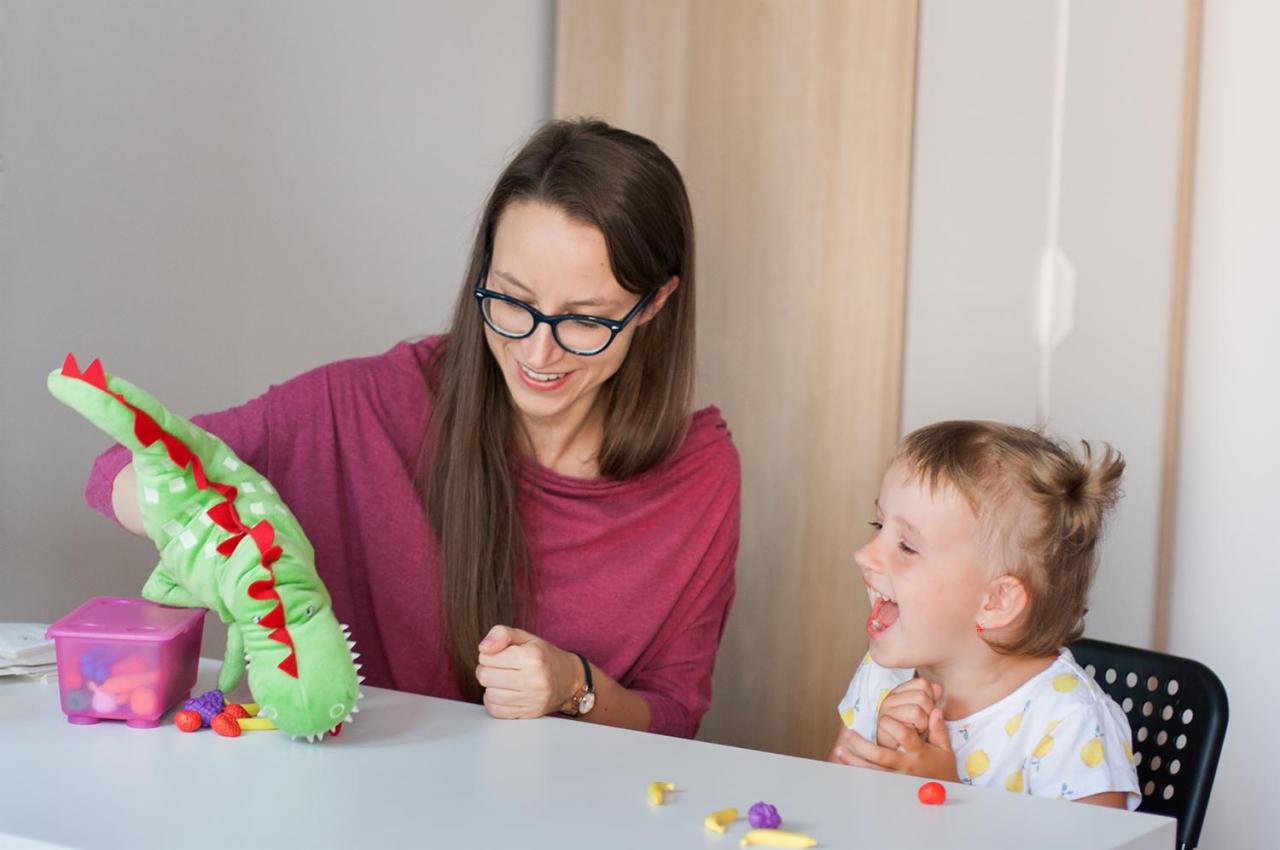 Logopeda prezentuje pacjentce pluszowego dinozaura z czerwonymi kolcami podczas terapii, na stole kolorowe plastelinowe kształty i pudełko z zabawkami.