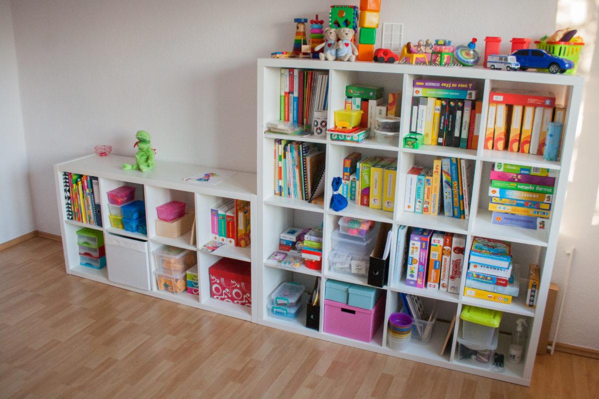 Dwie białe, modułowe półki wypełnione książkami, zabawkami i pojemnikami na zabawki, stojące na drewnianej podłodze, z pluszowym dinozaurem na górnej półce.