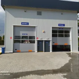 Szara hala z dwiema bramami garażowymi segmentowymi z oknami, oznaczonymi 'Brama 2' i 'Brama 3' z napisami 'Wyjazd' i 'Wjazd', widoczne znaki zakazu wjazdu, teren wyłożony kostką brukową.
