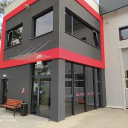 Nowoczesny budynek usługowy z szarymi ścianami i czerwonymi akcentami, szklanym zadaszeniem nad wejściem, widoczne okna i brama garażowa, ławka przed wejściem.