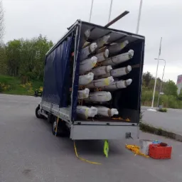 Ciężarówka z otwartą plandeką załadowana długimi, owiniętymi w folię przedmiotami, zabezpieczonymi drewnianymi belkami, z widocznymi pasami mocującymi i skrzynkami na ziemi.