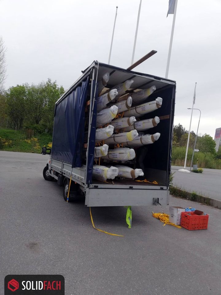 Ciężarówka z otwartą plandeką załadowana długimi, owiniętymi w folię przedmiotami, zabezpieczonymi drewnianymi belkami, z widocznymi pasami mocującymi i skrzynkami na ziemi.