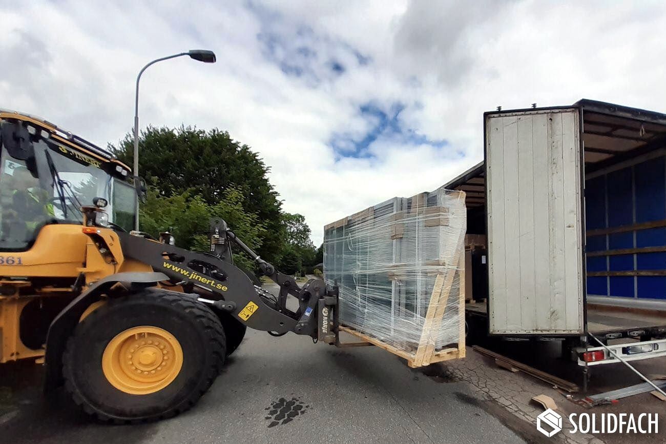 Żółta ładowarka Volvo podnosi paletę z oknami zabezpieczonymi folią, przygotowując je do załadunku na białą ciężarówkę z otwartą naczepą na tle zieleni i pochmurnego nieba.