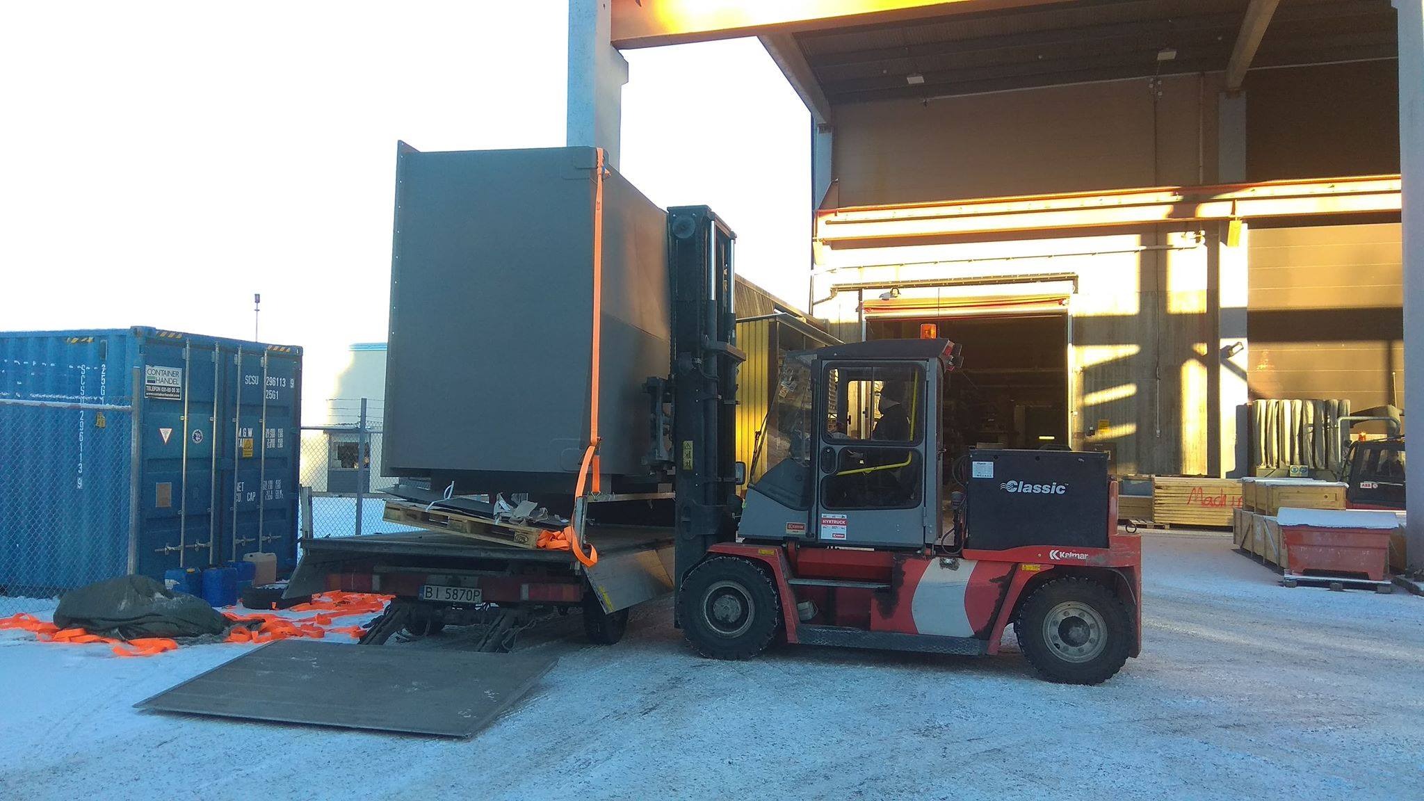 Wózek widłowy Kolmar transportuje duży, szary kontener na paletach, zabezpieczony pomarańczowymi pasami, z naczepy ciężarówki; w tle magazyn i kontenery transportowe.