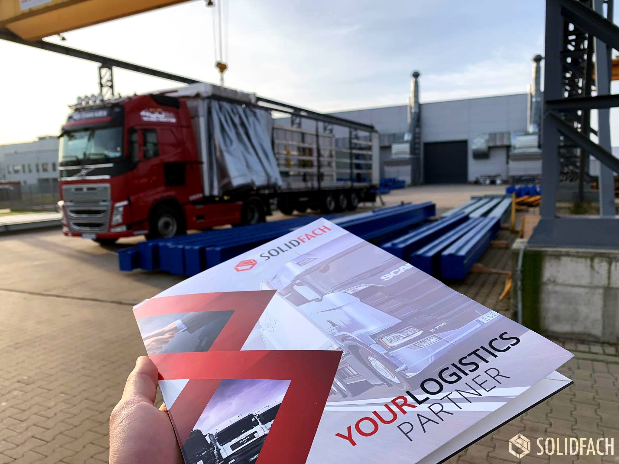 Broszura reklamowa firmy logistycznej Solidfach trzymana w dłoni na tle czerwonej ciężarówki z naczepą pod suwnicą.