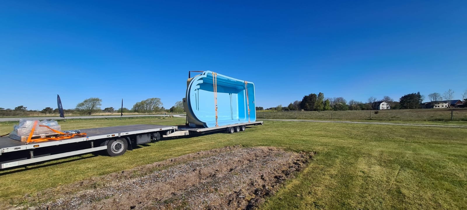 Niebieski basen z tworzywa sztucznego zabezpieczony pasami na platformie niskopodwoziowej, w tle zielone pole i błękitne niebo.