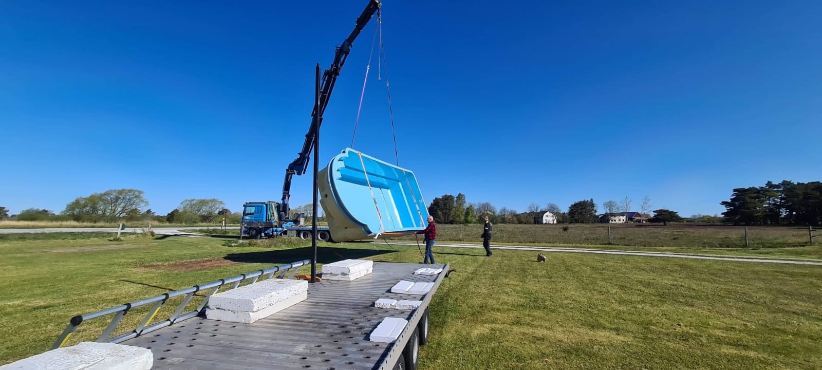 Kran podnosi prefabrykowany basen w kolorze błękitnym nad platformą transportową, w tle pracownicy i krajobraz wiejski.